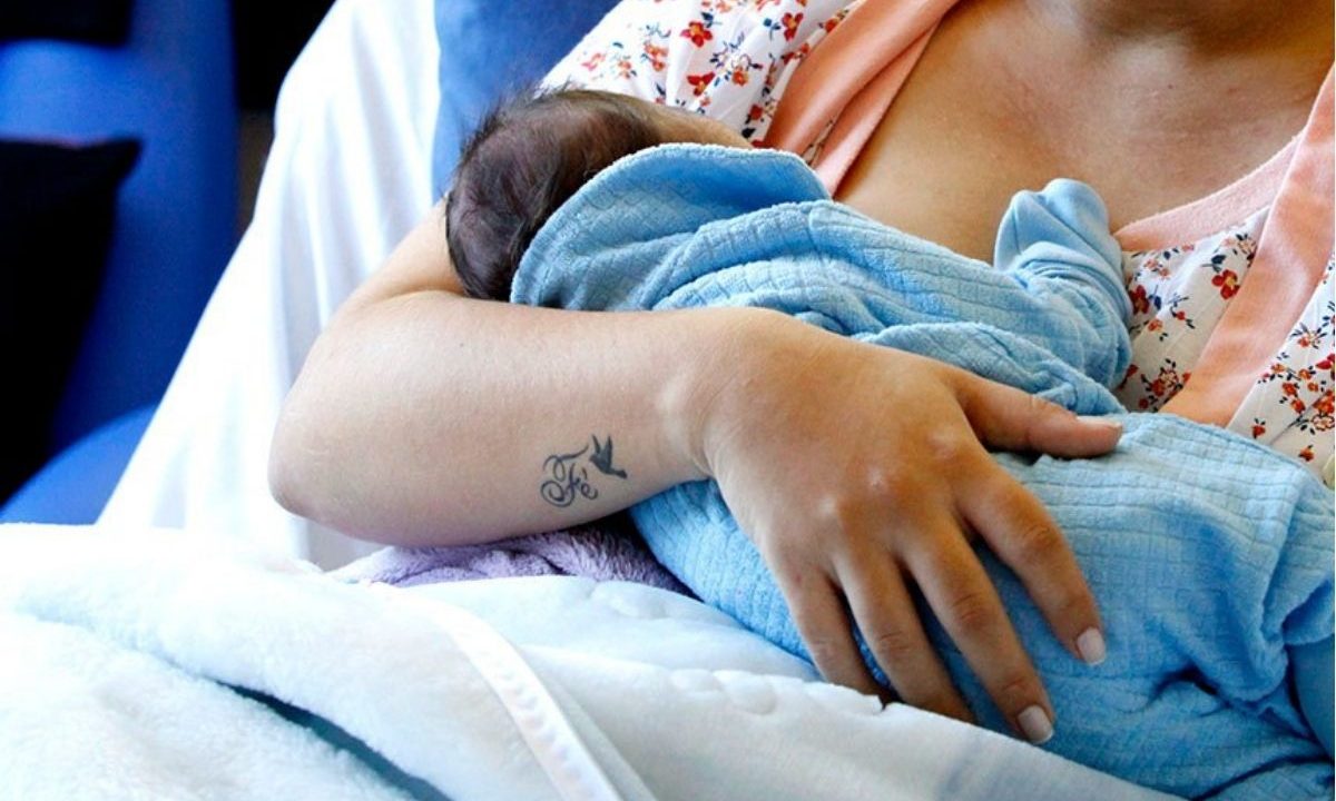 Mãe em leito de hospital segurando e amamentando o bebê recém-nascido envolto em uma manta azul, ilustrando o direito ao salário-maternidade.