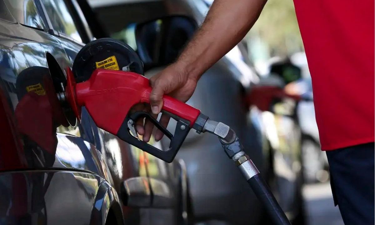 Frentista segurando bico de bomba de combustível vermelha e abastecendo tanque de carro preto em posto, ilustrando a medida de subsídio preço gasolina.