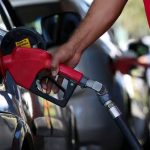 Frentista segurando bico de bomba de combustível vermelha e abastecendo tanque de carro preto em posto, ilustrando a medida de subsídio preço gasolina.