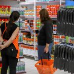 Cliente com cesta de compras sendo atendida por funcionária em corredor de loja de tintas e materiais de construção, ilustrando a discussão sobre os impactos no comércio com o fim da escala 6x1.