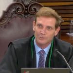 Plano médio do conselheiro Fábio Camargo sentado em uma cadeira alta de couro marrom escuro com detalhes entalhados em madeira, típica de um tribunal. Ele tem pele clara, cabelos loiros curtos, veste uma camisa social azul clara, gravata listrada em tons de roxo e cinza, e usa a toga preta de conselheiro com cordão verde. Ele está falando em direção ao microfone fixado à sua frente, com um notebook parcialmente visível na borda inferior.