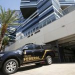Viatura preta da Polícia Federal estacionada em frente a um edifício moderno de arquitetura espelhada, ilustrando a operação contra fraude no INSS.