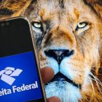 Smartphone exibindo o logotipo azul do aplicativo da Receita Federal em primeiro plano, com a imagem da cabeça de um leão ao fundo, ilustrando o prazo do imposto de renda.