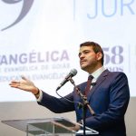 Fotografia em ângulo contra-plongé mostrando o ministro Jorge Messias, de perfil para a esquerda, discursando em um púlpito acrílico transparente. Ele tem pele clara, cabelos e barba escuros bem aparados, veste terno azul-escuro, camisa branca e gravata vinho. Seu braço esquerdo está estendido para a frente com a mão aberta gesticulando. Ao fundo, há um grande telão branco com inscrições parciais que incluem "UniEVANGÉLICA", "UNIVERSIDADE EVANGÉLICA DE GOIÁS" e a palavra "DIREITO".