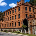 Vista externa da antiga fábrica fundada por Dom Pedro II construída com tijolos laranjas. Ao fundo, um céu bem azul, poucas nuvens e vegetação ao redor