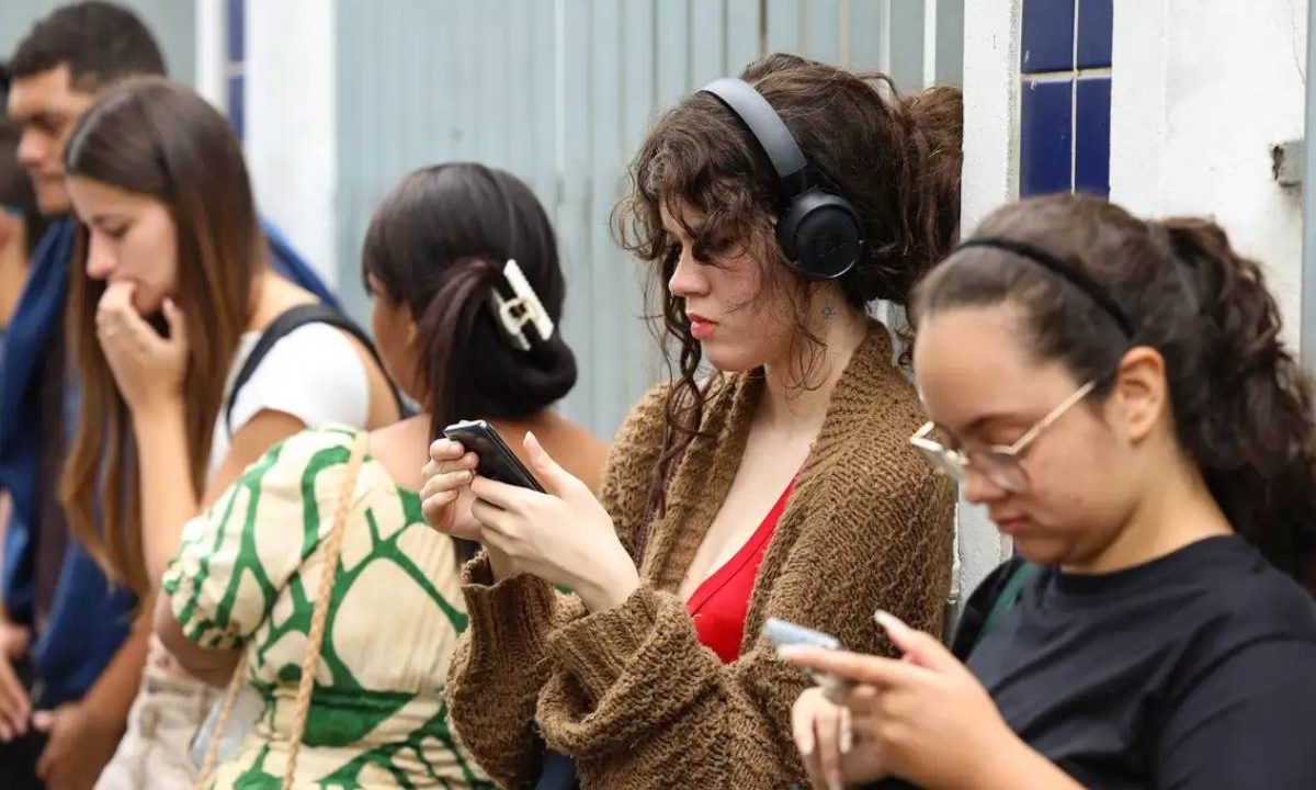 Jovens mexendo no celular enquanto aguardam em uma fila. Imagem ilustrativa para matéria sobre o novo Desenrola Fies que beneficia estudantes inadimplentes