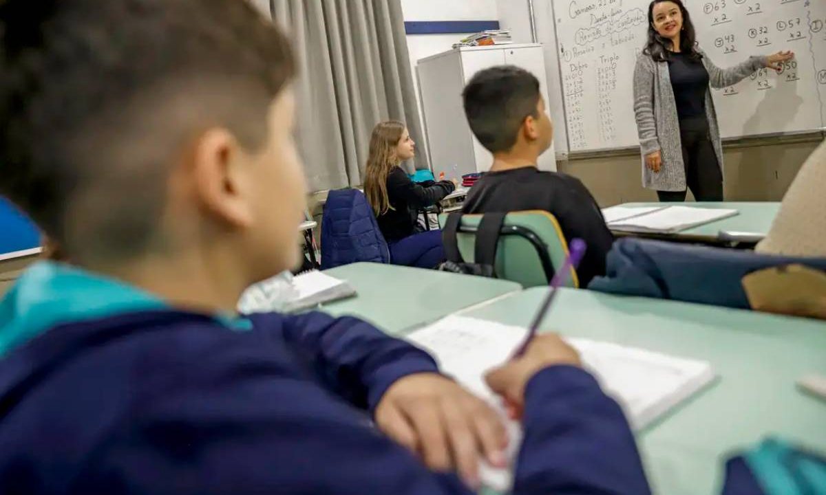 Criança escrevendo em seu caderno em sala de aula, ao fundo, professora lecionando. Imagem ilustrativa para matéria: Municípios do Paraná correm risco de perder recursos do Fundeb por pendências em dados obrigatórios