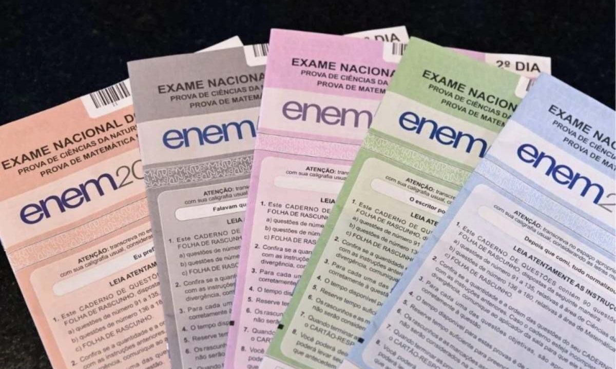 Mão segurando quatro cadernos de questões do Enem de cores diferentes (azul, amarelo, branco e verde). Imagem ilustrativa para matéria: Inep abre seleção para o Enem e pagarão até R$ 864 por dia; veja quem pode participar