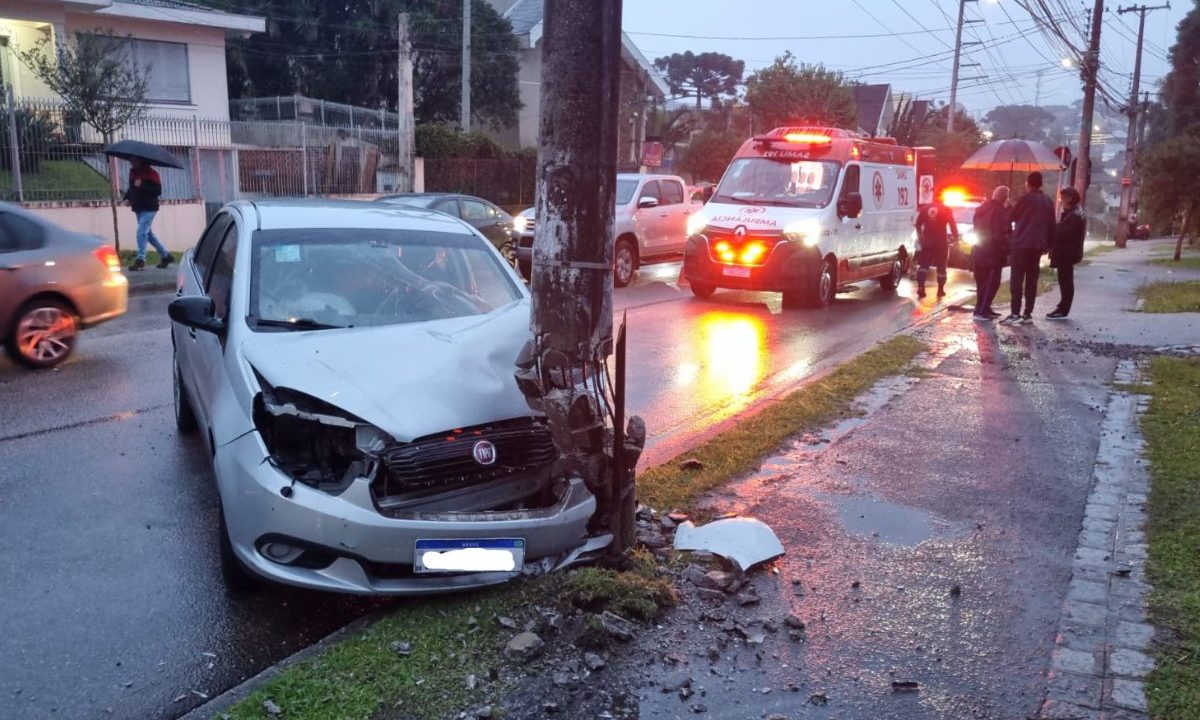 Na imagem aparece um carro modelo modelo Fiat Grand Siena após colidir contra um poste que teve a estrutura comprometida. Ao fundo a ambulância do Samu e outros veículos. O tempo nublado e chovendo. Idoso-apagão-perdeu-controle-direção-colisão-poste 1