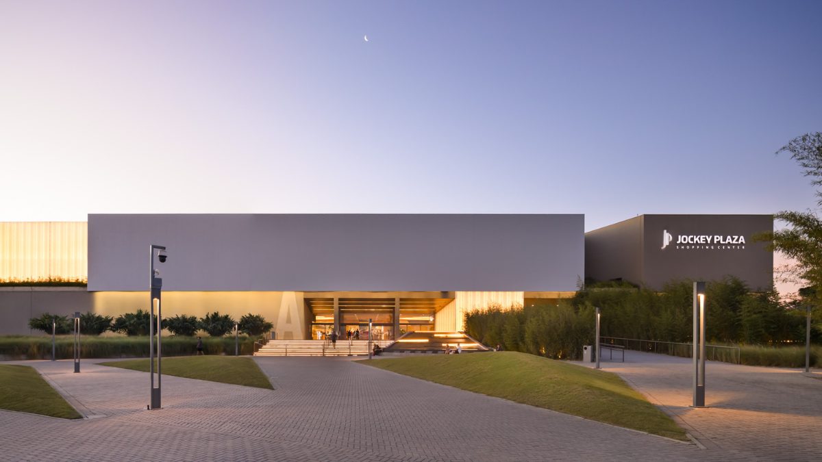 Fachada externa e moderna do Jockey Plaza Shopping iluminada ao anoitecer, com gramados e caminhos pavimentados em destaque.