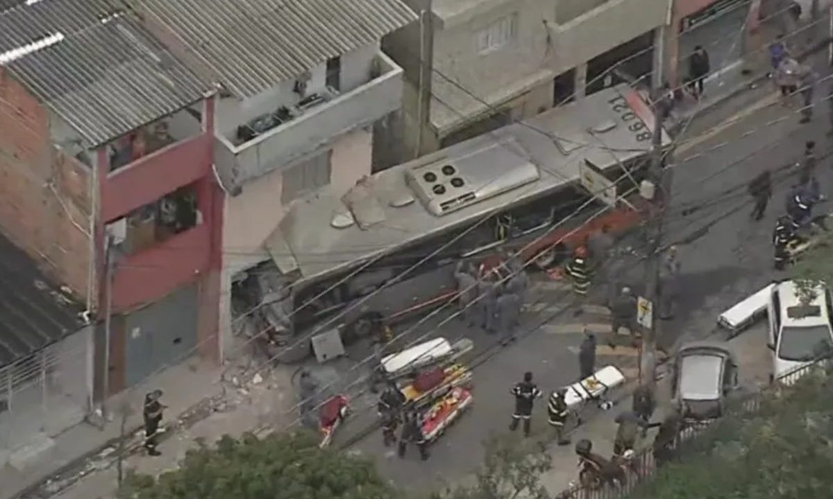 Uma imagem área feita por helicóptero mostra o ônibus atravessado n9o meio da via. Com a parte frontal dentro da residência. Autoridades na rua e cerca de 5 macas para atendimento médico