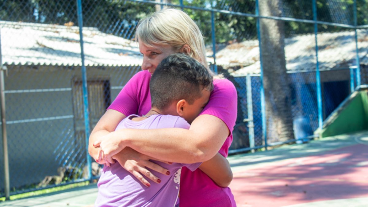 Mulher abraça criança em quadra esportiva durante ação do Programa Família Acolhedora em Curitiba.