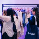 Uma cliente de óculos e cabelos longos segura uma grande sacola jeans da Shein enquanto observa peças de roupa coloridas nas araras em plano médio, ilustrando o movimento na Loja da Shein pop-up.