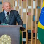 O presidente Luiz Inácio Lula da Silva fala ao microfone em um púlpito de madeira com o brasão da República, tendo ao fundo a bandeira nacional, ilustrando suas críticas às declarações vindas dos EUA.