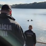 Dois policiais civis de costas, vestindo jaquetas escuras com a inscrição "Polícia Civil", observam mergulhadores do Corpo de Bombeiros na água durante operação para busca de armas encontradas represa.