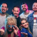 Os integrantes da banda DeLorean posam juntos sorrindo em um ambiente externo com árvores ao fundo, ilustrando a divulgação do show de aniversário.