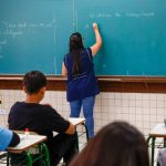 Professora de costas escrevendo "História da computação" com giz no quadro verde enquanto alunos assistem à aula, ilustrando a aprovação do piso professores.