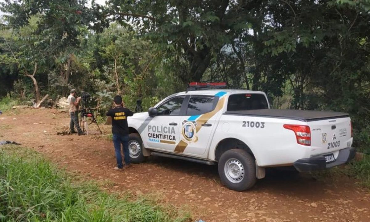 Na imagem ilustrativa mostra uma viatura da Polícia Científica do Paraná e dois homens, em uma estrada de chão, repleta de vegetação.