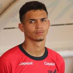 Arthur Dias aparece de pé, usando camisa vermelha de treino do Athletico, em registro próximo ao banco de reservas. O zagueiro observa o campo com expressão séria durante atividade do clube.