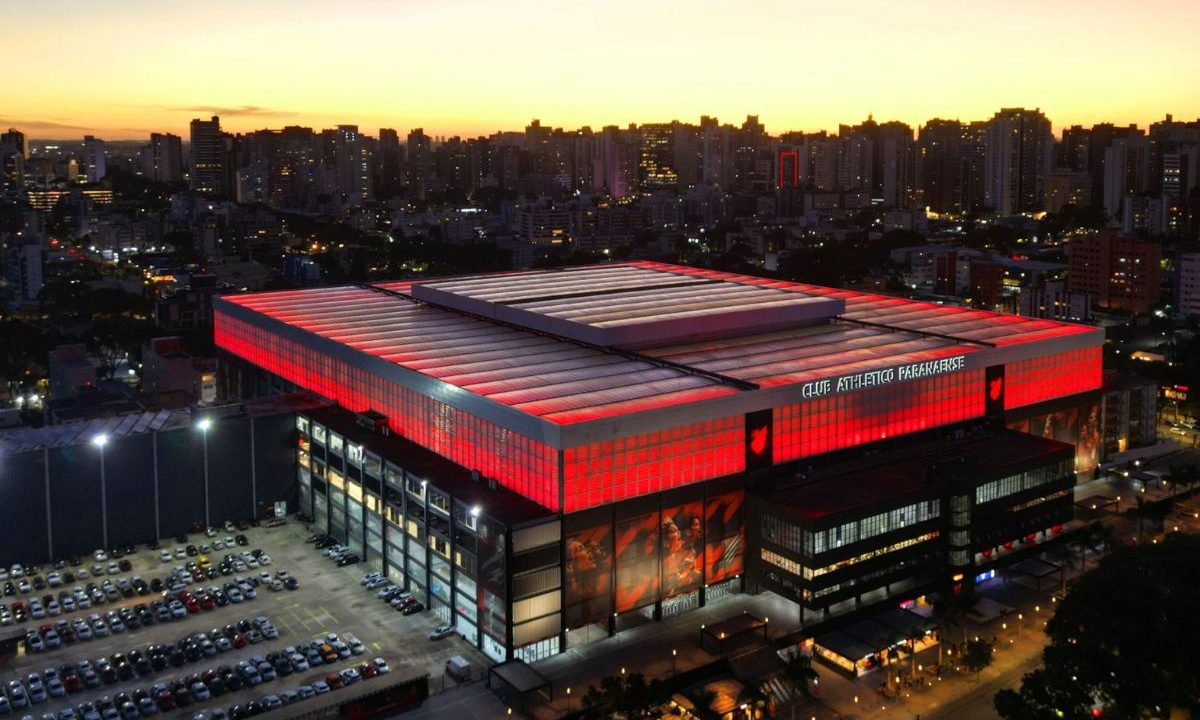 Imagem aérea mostra a Arena do Athletico iluminada em vermelho no início da noite, cercada pelos prédios da cidade de Curitiba. O estádio aparece com o teto fechado e destaque para a iluminação externa.