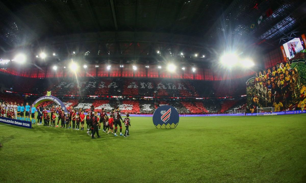 Visão ampla da Arena da Baixada durante entrada das equipes, com mosaico nas arquibancadas e bandeirão em homenagem a jogadores ligados ao Athletico e à Seleção Brasileira.