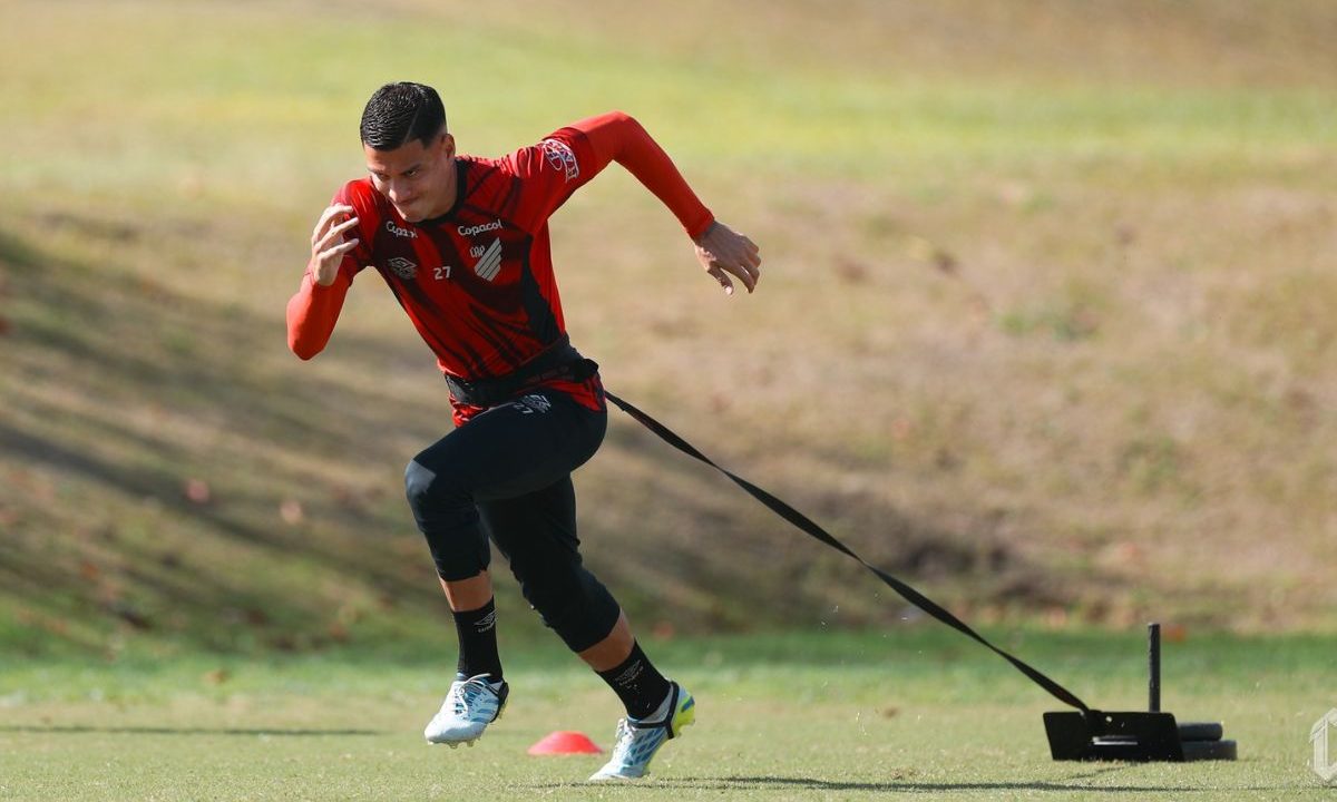 Jogador Juan Portilla aparece durante treinamento físico no CT do Athletico. O volante colombiano veste uniforme de treino rubro-negro e realiza exercício de arrancada com equipamento de resistência preso à cintura em campo aberto.