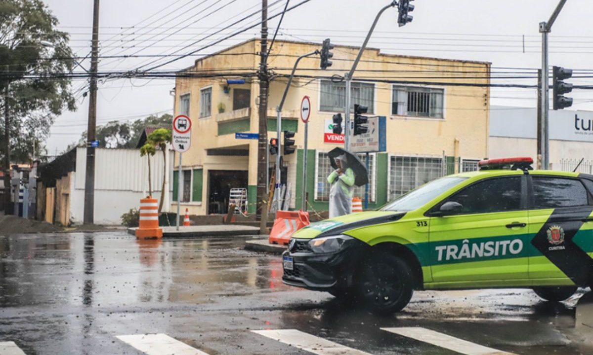 Viatura da Superintendência de Trânsito bloqueia cruzamento em Curitiba durante chuva, enquanto agentes organizam o fluxo de veículos na região.