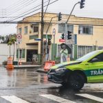 Viatura da Superintendência de Trânsito bloqueia cruzamento em Curitiba durante chuva, enquanto agentes organizam o fluxo de veículos na região.