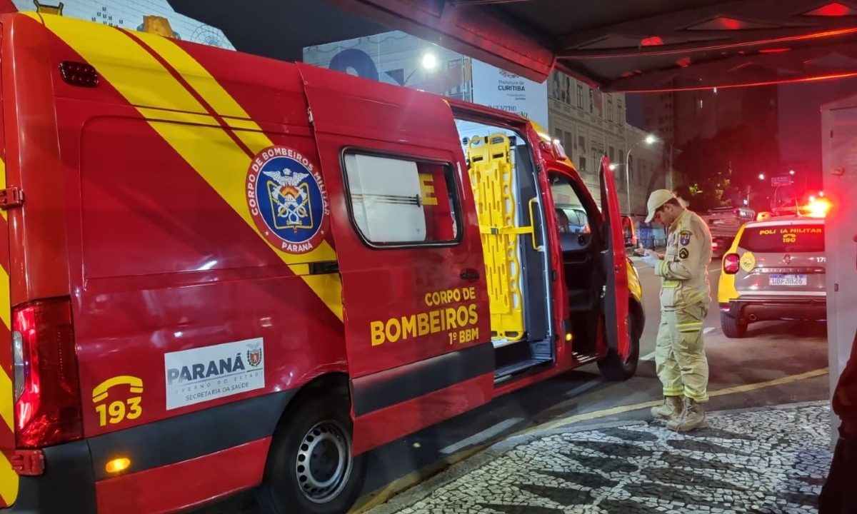 Foto tirada durante a noite mostra bombeiro ao lado de ambulância da corporação anotando informações em uma prancheta. A ambulância está parcialmente sobre uma calçada, a poucos metros de um ponto de ônibus. Socorristas atendem homem esfaqueado após briga de bar.