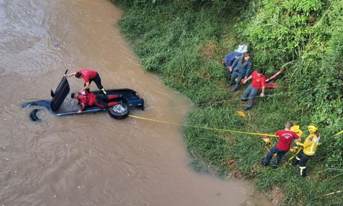 Foto mostra bombeiros resgatando carro que caiu em rio e mulher que morreu afogada presa ao cinto de segurança, em Santa Catarina. O carro está de lado, parcialmente submerso na água turva. Dois bombeiros estão sobre o veículo, que é puxado com uma corda. Outros bombeiros estão às margens auxiliando.