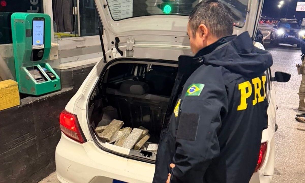 Na imagem aparece um agente da Polícia Rodoviária Federal parado ao lado do bagageiro do automóvel — modelo Volkswagen Gol, branco — aberto, com tabletes de maconha embalados, em um pedágio.