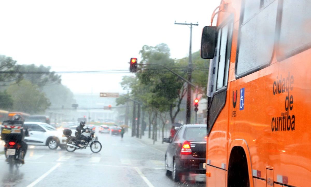 Uma rua de Curitiba em um dia chuvoso e nublado em frente fria no Paraná. Em primeiro plano, à direita, a lateral de um ônibus laranja da frota urbana, com a inscrição "cidade de curitiba" e o símbolo de acessibilidade. No centro, o asfalto molhado reflete as luzes dos veículos. Há carros cinzas e motos circulando sob a chuva, além de semáforos com a luz vermelha acesa e árvores ao fundo.