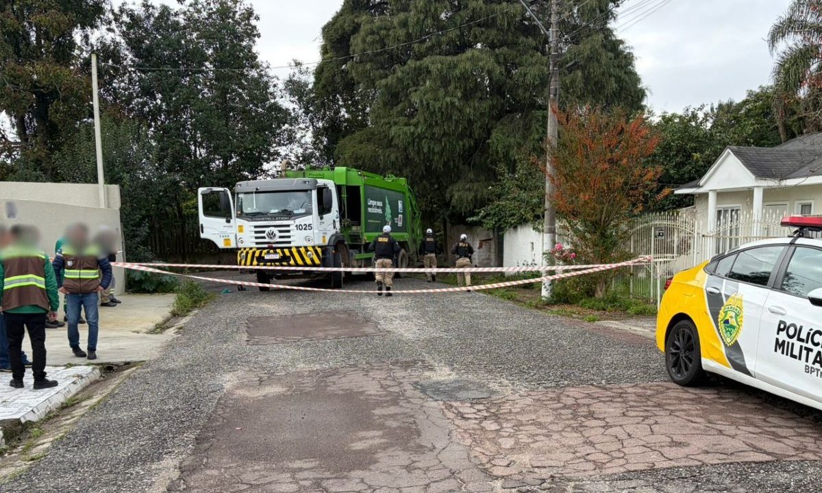 Caminhão de coleta de lixo, policiais e funcionários em rua no bairro Cajuru, em Curitiba, onde coletor de lixo morreu ao ser prensado contra muro.