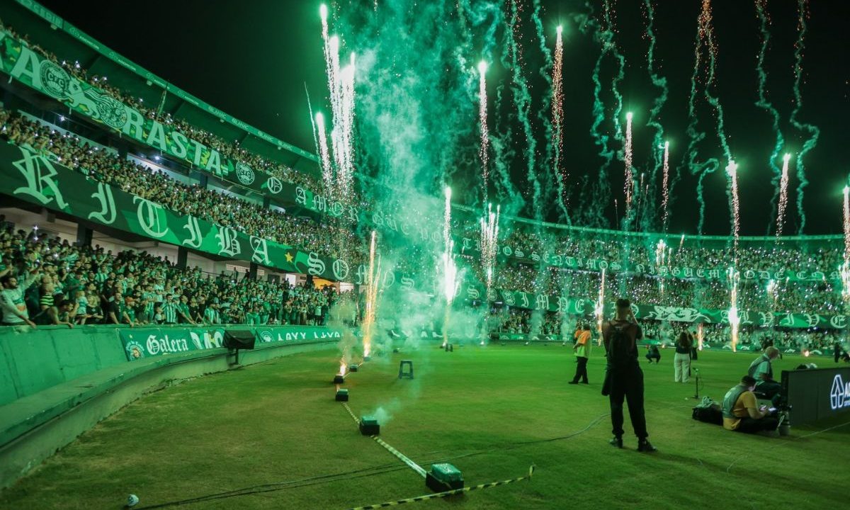 Imagem mostra o Couto Pereira lotado com a torcida do Coritiba fazendo festa nas arquibancadas. Fogos de artifício iluminam o gramado e o estádio aparece tomado pelas cores verde e branca antes da partida.