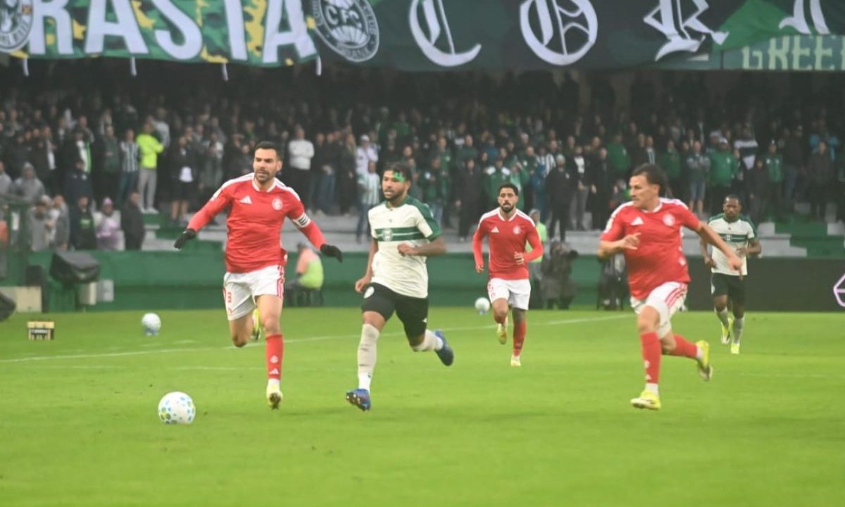 Imagem mostra um jogador do Coritiba conduzindo a bola em velocidade no gramado do Couto Pereira durante partida contra o Internacional. Atletas colorados aparecem tentando acompanhar a jogada, enquanto torcedores alviverdes ocupam as arquibancadas ao fundo com bandeiras e faixas do clube.