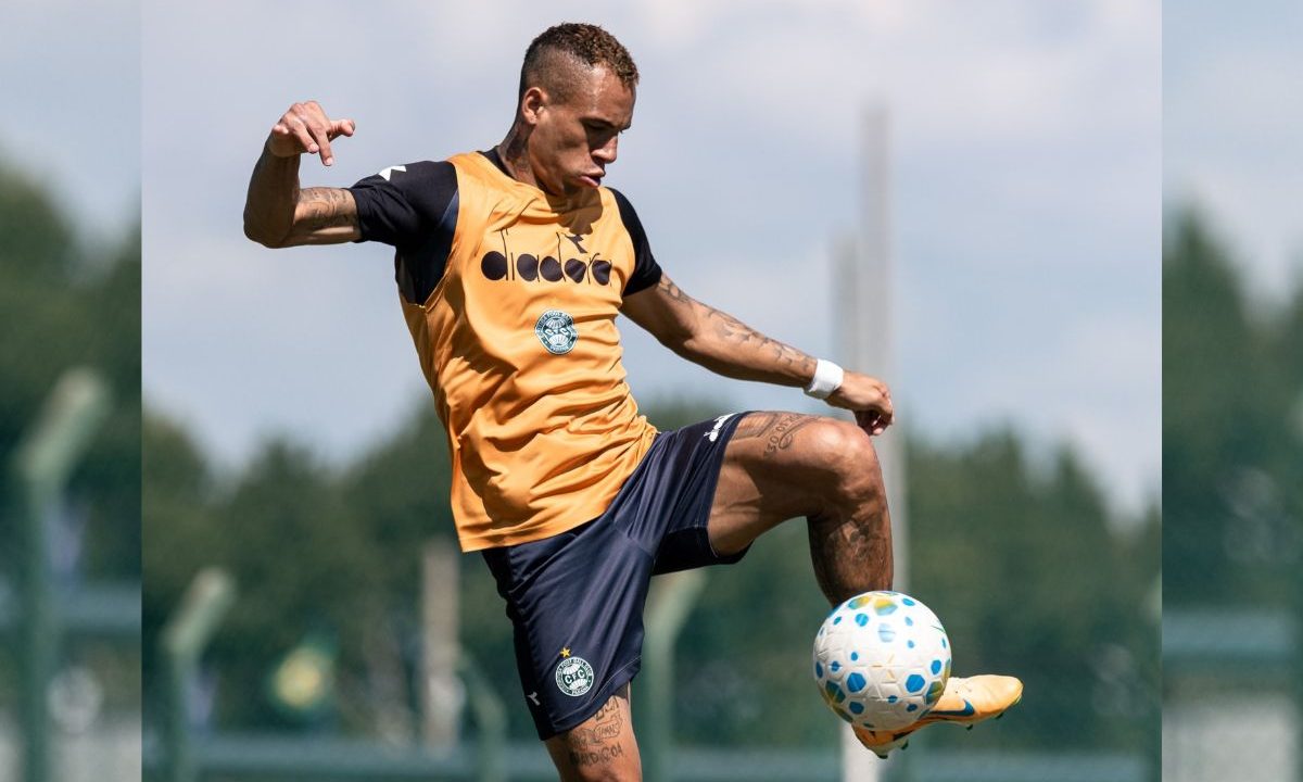 Breno Lopes aparece durante treinamento do Coritiba no CT da Graciosa. O atacante domina a bola com a perna direita enquanto realiza atividade em campo aberto. O jogador usa colete amarelo sobre o uniforme de treino alviverde. Ao fundo, o gramado e a paisagem aparecem desfocados.
