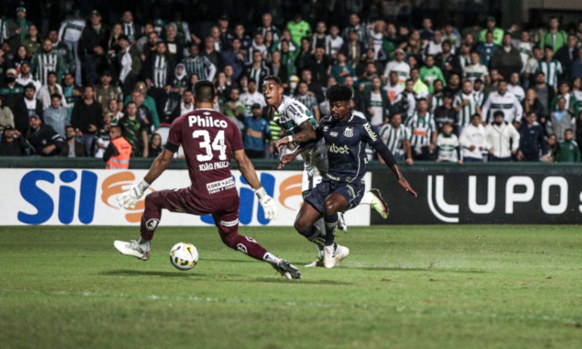 A imagem mostra o atacante Alef Manga, do Coritiba, no momento da finalização que resultou no gol da vitória sobre o Santos pela Copa do Brasil 2022. O goleiro santista tenta fechar o ângulo enquanto torcedores acompanham o lance ao fundo, no Couto Pereira.