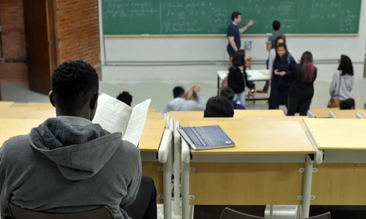 estudante negro sentado em um auditório universitário, lendo um material, enquanto outros alunos e um professor aparecem ao fundo em uma sala de aula. Imagem ilustrativa para matéria: Fraude em cota racial leva estudante de medicina a pagar multa de R$ 720 mil