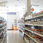 Interior de farmácia com prateleiras abastecidas de medicamentos, produtos de higiene e balcão de atendimento ao fundo.