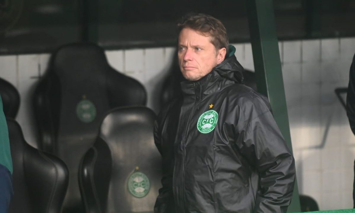 Imagem mostra o técnico Fernando Seabra na área técnica do Couto Pereira durante partida do Coritiba. O treinador veste casaco preto com o escudo do clube e observa o campo com expressão séria, enquanto os bancos de reservas aparecem ao fundo.