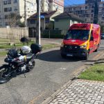 Imagem mostra uma rua em dia com céu azul. Há prédios. Um carro está estacionado no lado direito da rua. Uma motocicleta da polícia militar está no meio da via e uma ambulância do Siate estacionada em atendimento ao homem que foi contido com arma de choque no Juvevê. Dois policiais estão na calçada conversando.