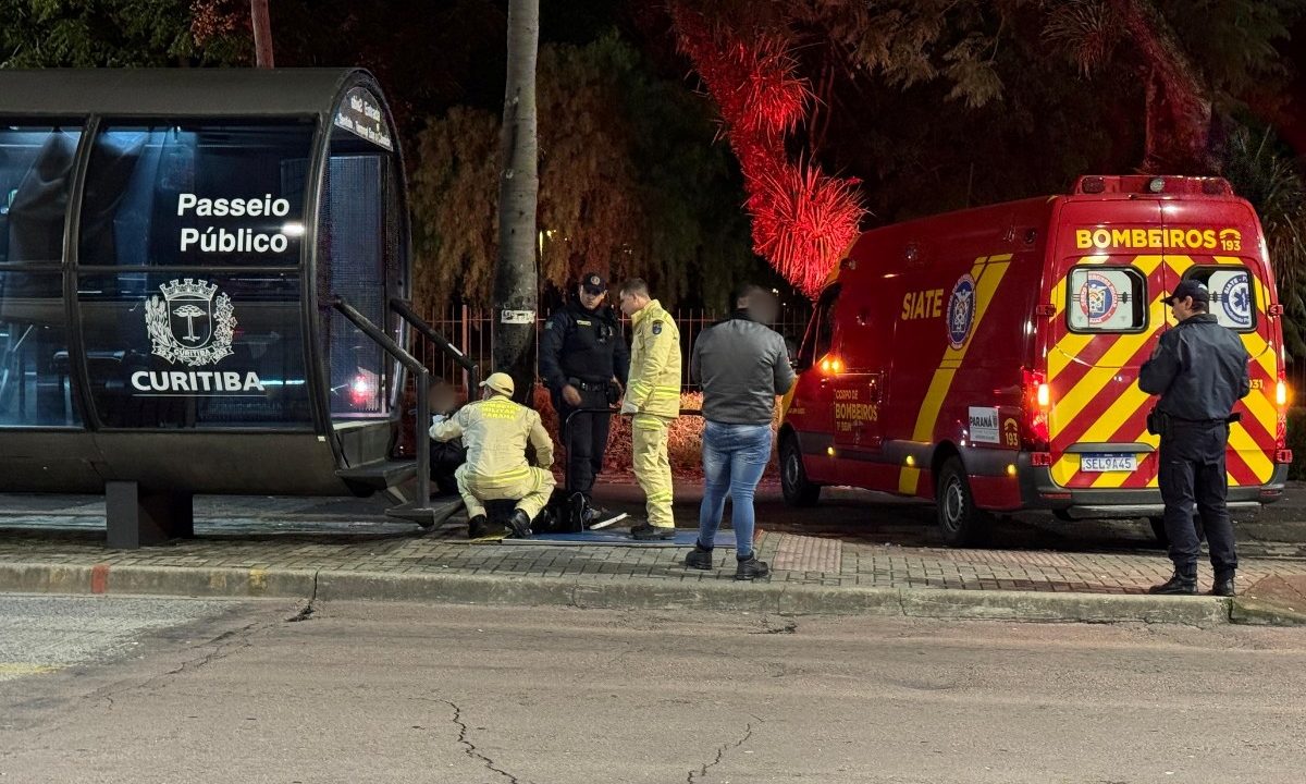 Foto tirada à noite, de longe, mostra homem confundido com torcedor que foi espancado recebendo ajuda de bombeiro, policial militar e guarda municipal ao lado de estação-tubo na região do Passeio Público, em Curitiba