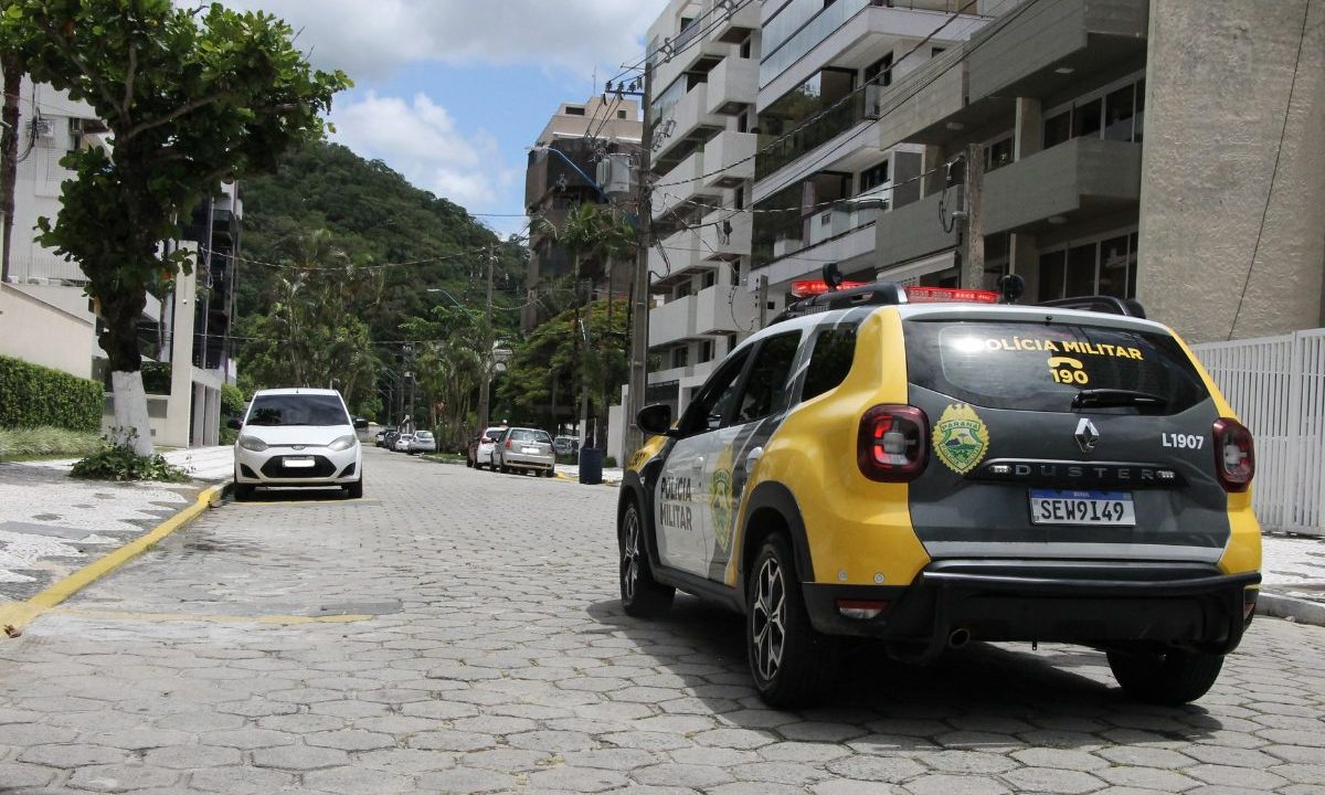 Na imagem, uma viatura da Polícia Militar aparece estacionada em uma rua de paralelepípedos. No sentido contrário, há outros veículos. Ao fundo, uma montanha coberta por árvores se destaca na paisagem, cercada por prédios às margens da via