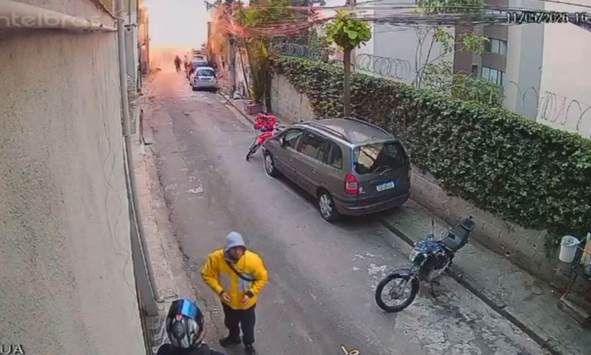 Captura de uma câmera de segurança que registrou o momento da explosão. A imagem mostra uma rua estreita, com alguns veículos estacionados na lateral da via, um clarão ao fundo ocasionado pela explosão e dois homens conversando próximos a câmera, que está distante do incidente. homem-morre-pessoas-feridas-casa-explodir