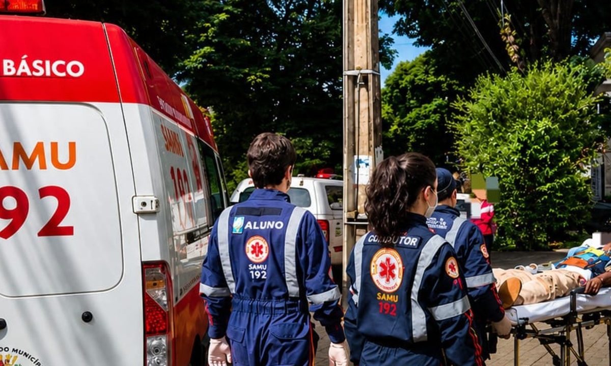 Foto mostra socorristas do Samu caminhando e transportando em cima de uma maca homem que foi atacado a facadas ao sair para comprar presente de Dia das Mães. Pelo menos quatro socorristas atuam no socorro. Uma ambulância do Samu aparece estacionada em frente ao que parece ser um hospital