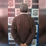Na imagem aparece o idoso posando de costas ,algemado, em uma delegacia da Polícia Civil