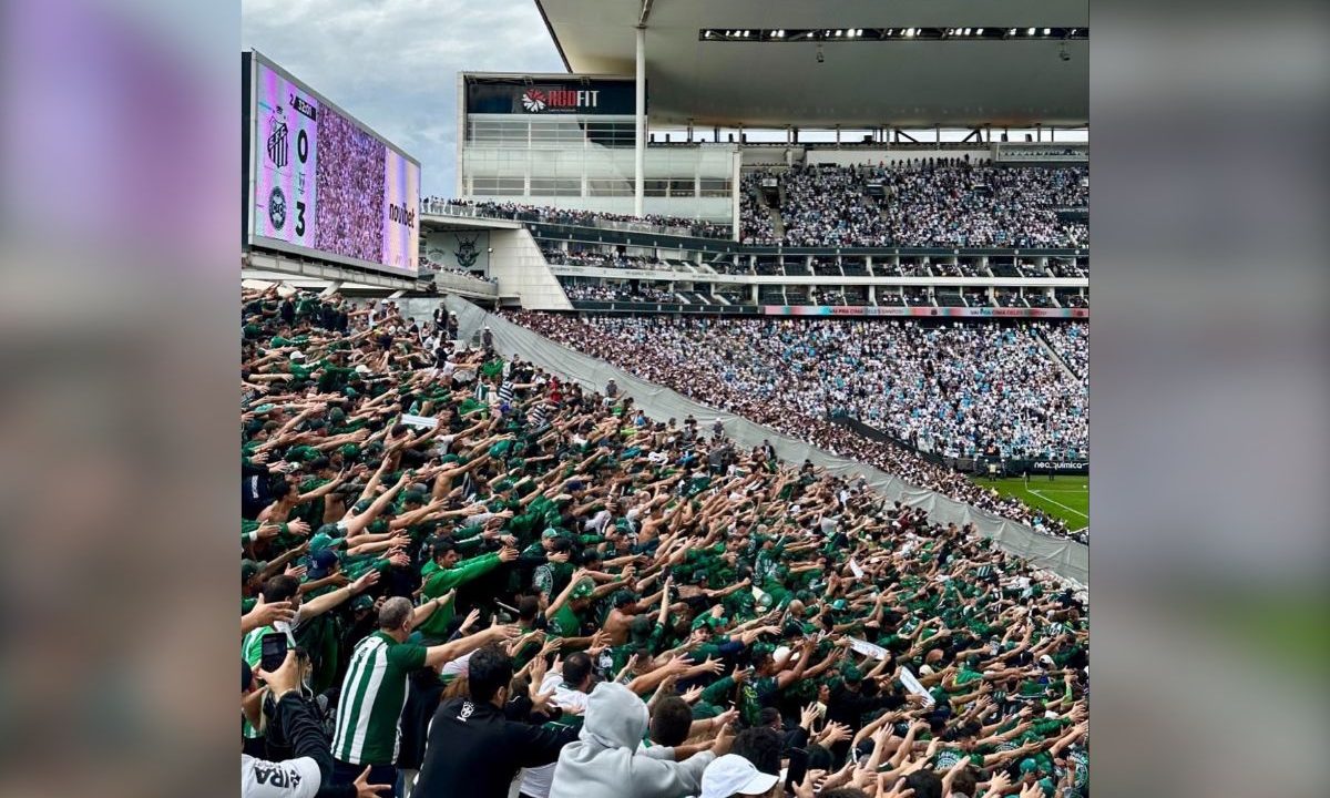 Membros da Império Alviverde, principal organizada do Coritiba ocupam setor da Neo Química Arena com os braços erguidos em apoio ao time. Ao fundo, o telão do estádio exibe o placar de 3x0 para o Coxa sobre o Santos