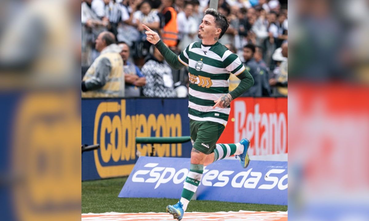 A imagem mostra Josué comemorando durante uma partida do Coritiba. O meia aparece correndo e apontando para o alto com o uniforme alviverde, enquanto a torcida acompanha o momento ao fundo nas arquibancadas