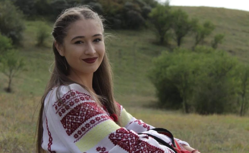 A jovem cantora de 17 anos morreu após sofrer um AVC. Minutos antes de subir ao palco, ela sentiu fortes dores de cabeça. Duas semanas após o AVC, ela morreu devido ao acidente vascular cerebral. Imagem mostra a jovem posando para a foto. Ela está sentada em um gramado verde, ela usa blusa colocirda com cores vinho e amarelo. Ela está com cabelo meio preso e batom forte escuro.
