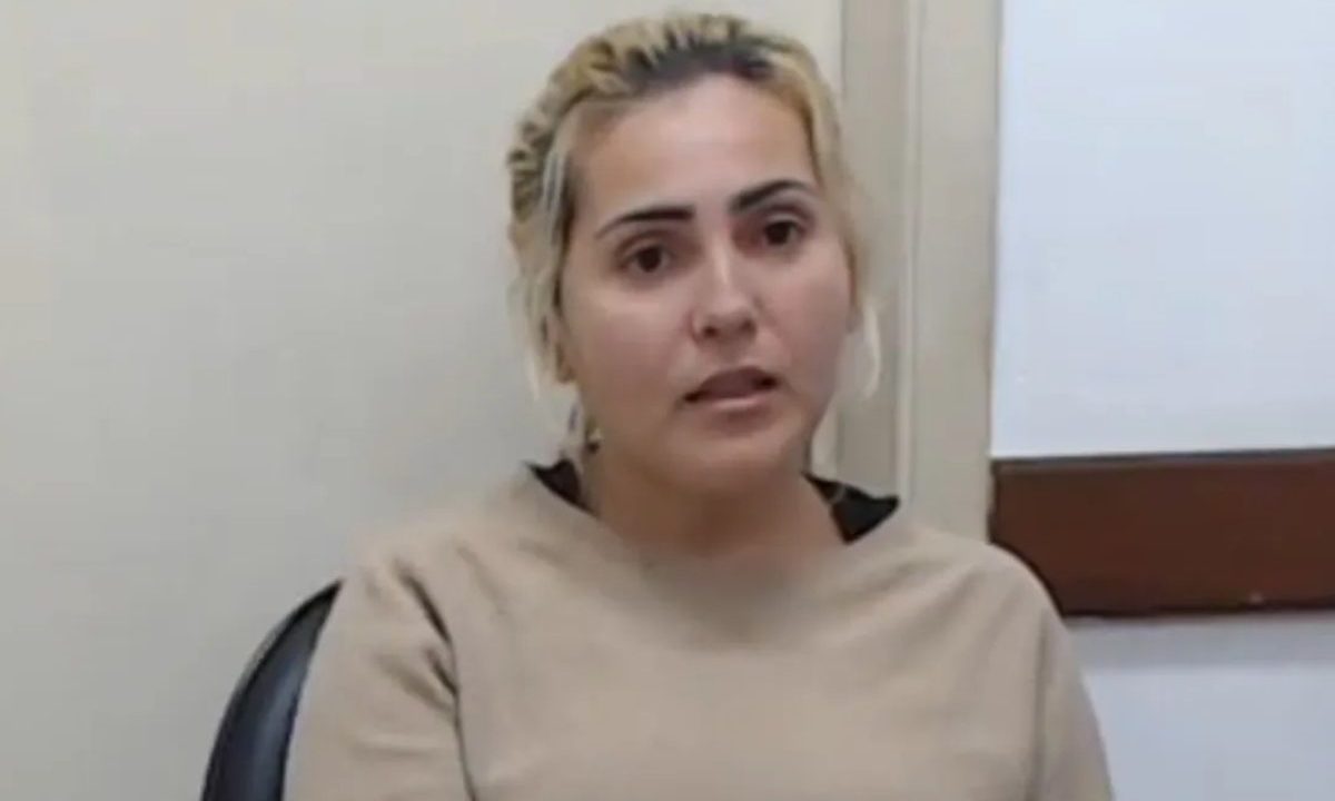 Na imagem aparece Ana Paula presa, aguardando julgamento, com o cabelo preso, uma camiseta marrom e sentada em uma sala simples